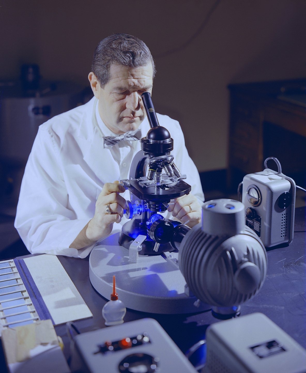Hombre con bata de laboratorio examina una muestra bajo un microscopio con luz azul.